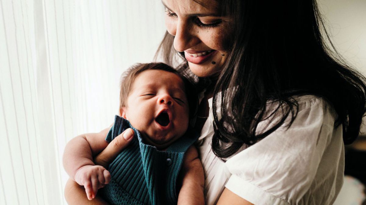 Eine junge Frau mit langen dunklen Haaren hält ein zufrieden gähnendes Baby im Arm.