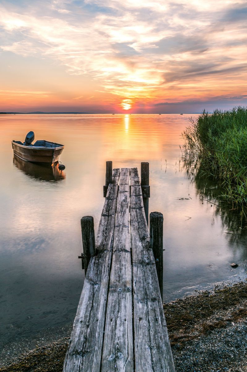 Sonnenuntergang über dem Bodensee, Blick von einem schmalen Holzsteg im Schilfgürtel, daneben ankert ein kleines hölzernes Motorboot im Wasser.