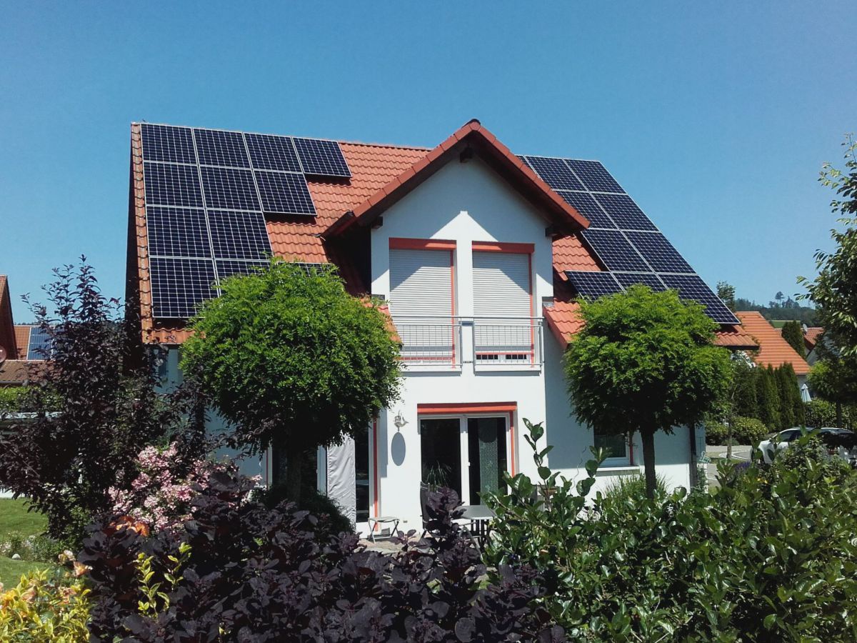 Das Bild zeigt ein Einfamilienhaus mit rotem Ziegeldach, das eine Photovoltaikanlage trägt. Rundherum eine Wohngegend mit viel Grün aus Gärten, Bäumen und Sträuchern und strahlend blauer Himmel.