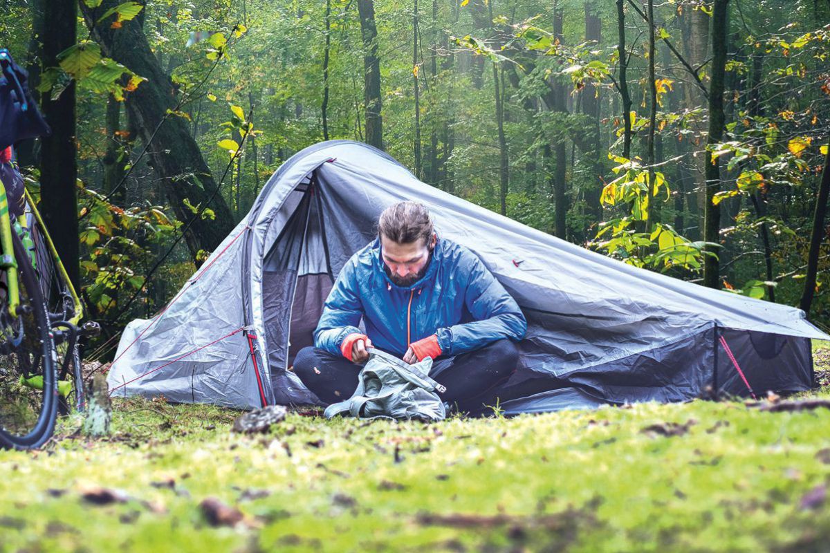 Auf einer kleinen LIchtung in einem Wald sitzt ein Mann in warmer Outdoorkleidung vor einem Zelt und wühlt in einem Stoffbeutel. Neben dem Zelt steht ein Fahrrad.