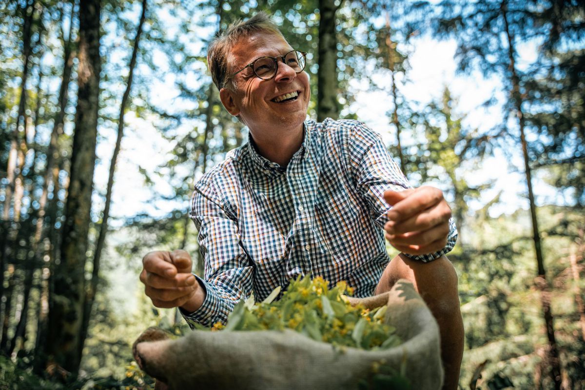 Ein Mann kniet im Wald neben einem Sack voller Lindenblüten. Er lacht.