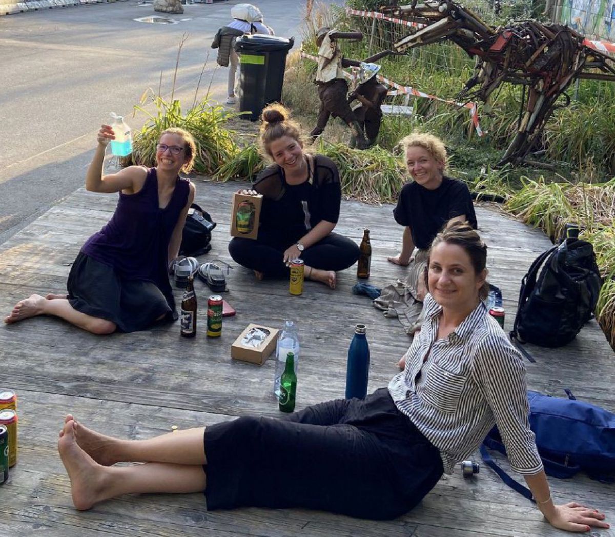 Eine kleine Gruppe Frauen sitzen zusammen auf einem Holzdeck am Ufer der Donau in Wien, es wirkt sommerlich warm, sie haben Getränkeflaschen bei sich und wirken fröhlich und entspannt.