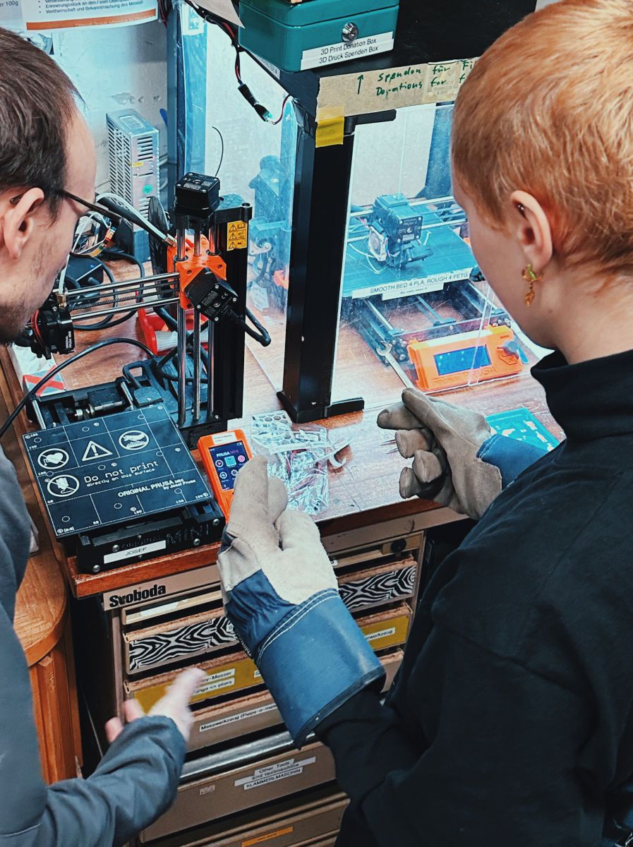 Zwei Personen sind an einem Werktisch mit Lasercutter und 3D-Drucker beschäftigt. Im linken Bildausschnitt ein Mann mit dunklen, kurzen Haaren und Brille, rechts eine junge Frau mit kurzen rötlichblonden Haaren, ihre Hände stecken in groben Arbeitshandsch