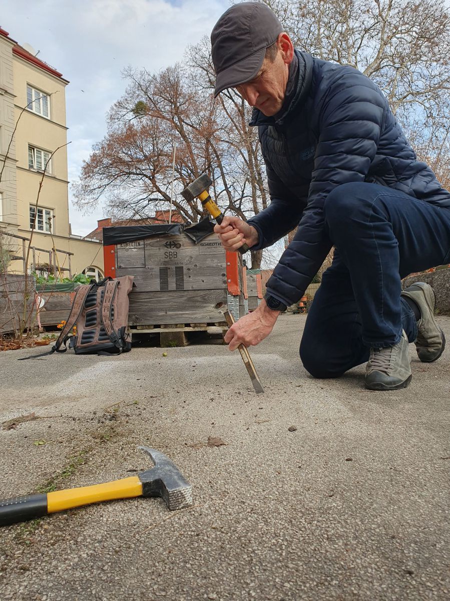 Ein Mann mit Schirmkappe, in dunkelblauen Jeans und Steppdaunenjacke, kniet auf der Fahrbahn in einer Wohnsiedlung und macht sich mit Hammer und Meißel daran, den Asphalt aufzubrechen. 