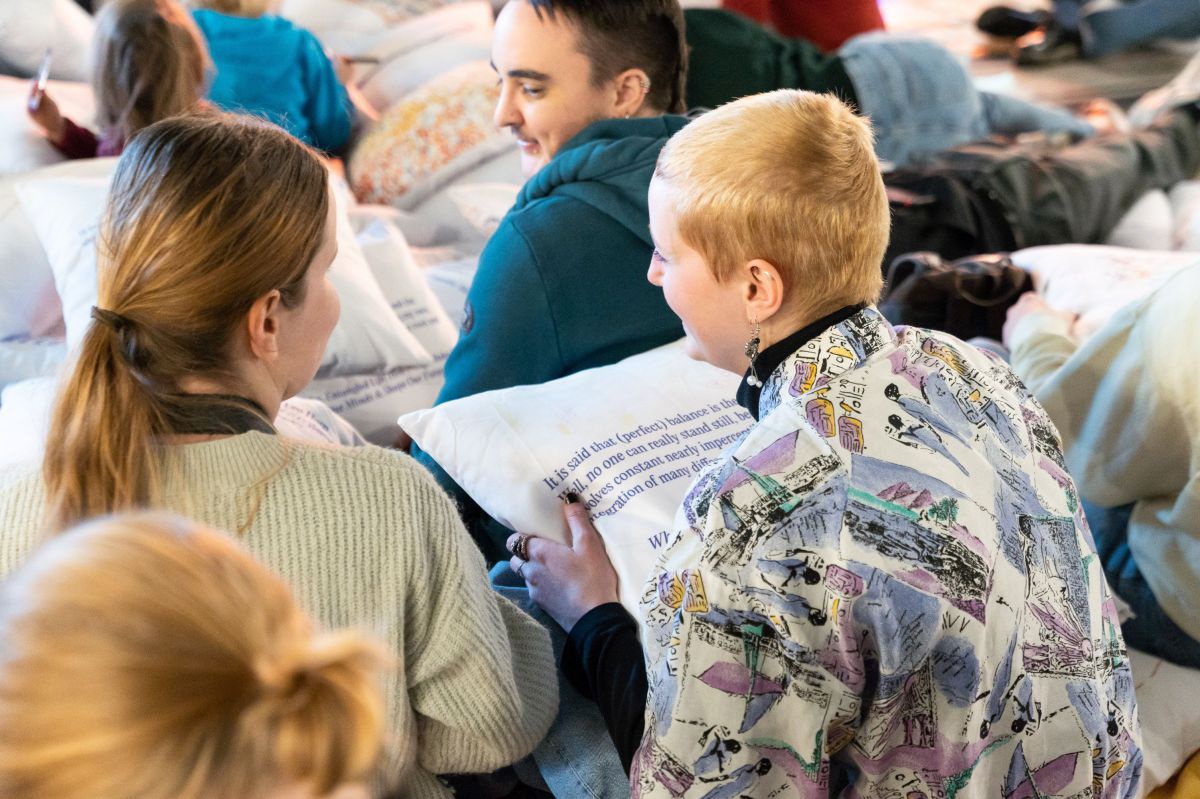 Menschen sitzen in Reihen am Boden, jede Person hält einen mit Text oder Illustrationen bedruckten Polster in Händen. Die Leute scheinen Gespräche miteinander zu führen.