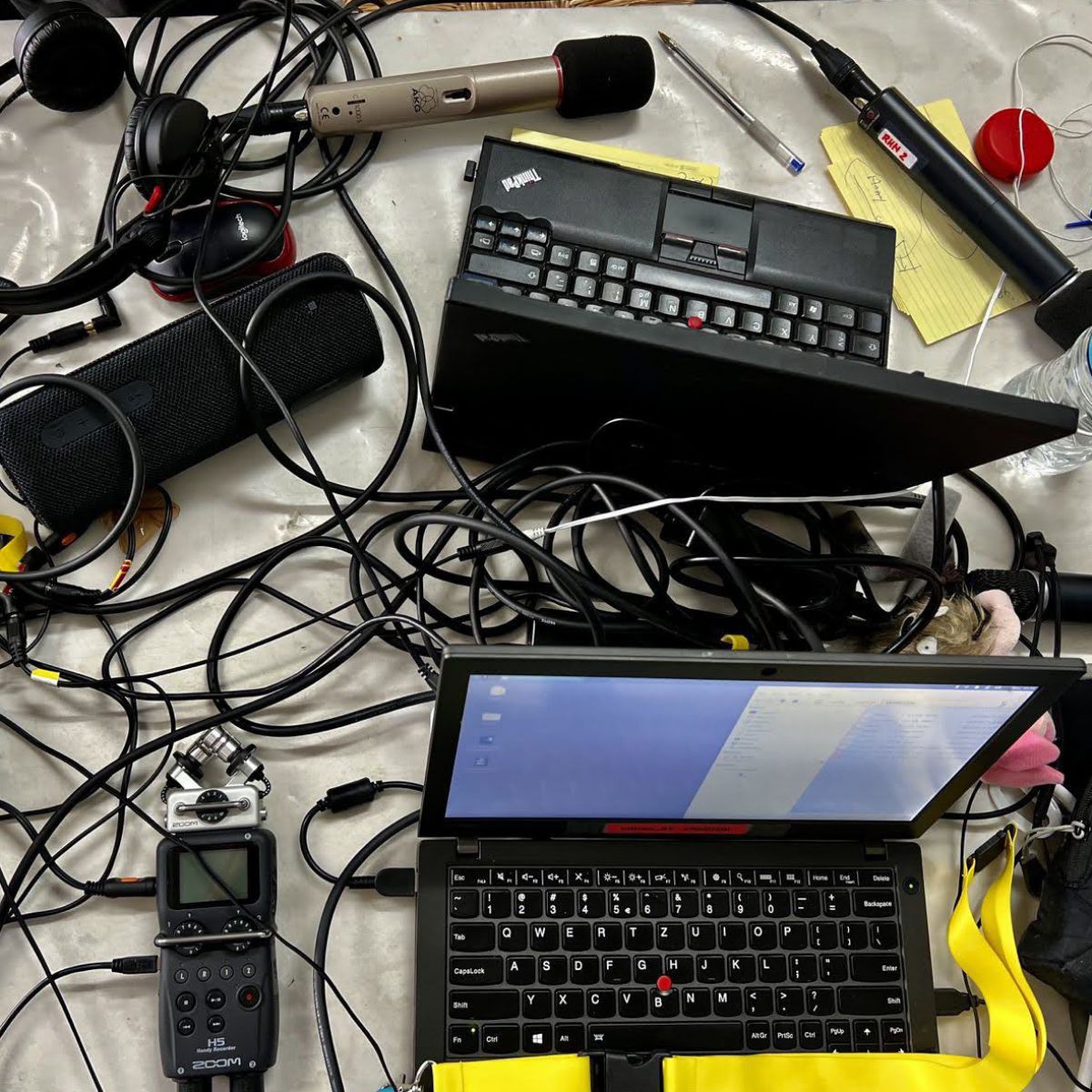 Radio-Equipment auf einem Tisch: Laptops, Mikrofone, Bluetoothlautsprecher, Kopfhörer, Aufnahmegerät, Kabeln, Notizzettel und Kuli, Wasserflasche.