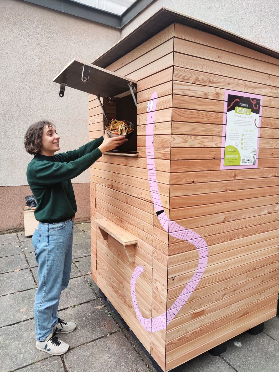 Eine junge Frau ist gerade dabei, Obst- und Gemüseabfälle in die geöffnete Klappe der Wurmkiste zu werfen. Auf dem hölzernen Container ist mit rosa Farbe ein Comic-Kompostwurm aufgemalt, auch ein Infoplakat ist an einer Wand befestigt.