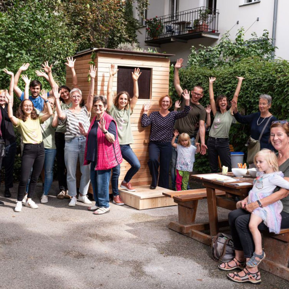 Um die 20 Menschen (Erwachsene und Kinder), haben sich um ein WurmHotel versammelt und posieren für das Foto. Die meisten winken und lachen fröhlich.