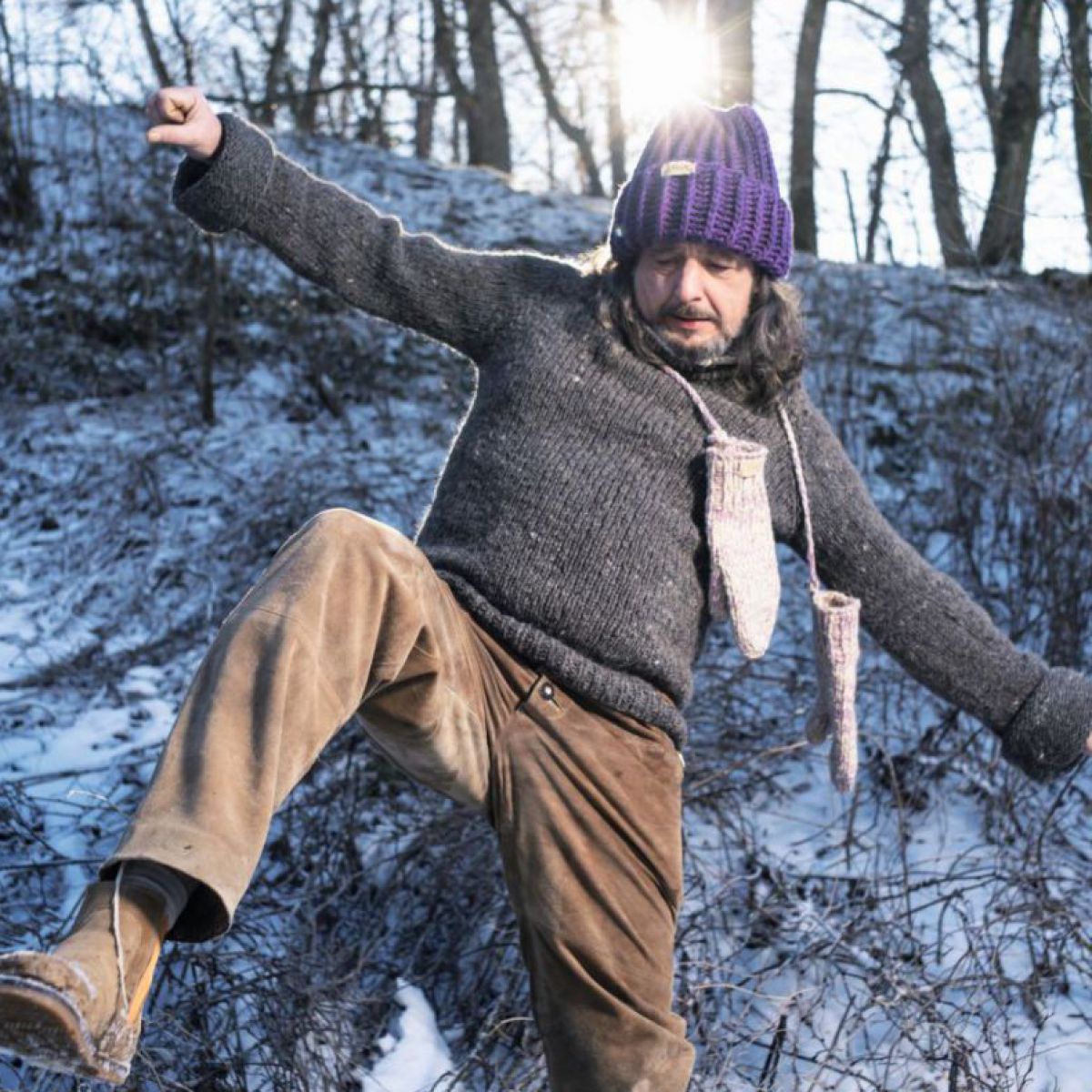 Ein Mann mit schulterlangem, lockigen Haar und Bart versucht auf dem gefrorenen Untergrund in einem  winterlichen Park das Gleichgewicht wieder zu erlangen. Er trägt einen grob gestrickten grauen Wollpullover, hat gestrickte naturfarbene Fäustlinge an 