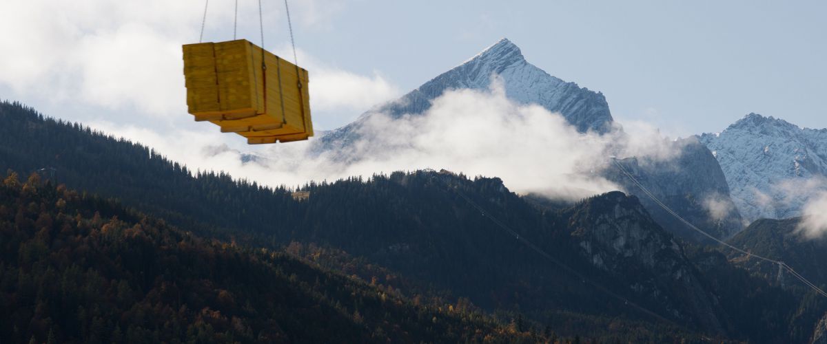 Ein Bergpanorame mit einer Palette Holz die durch einen Kran von einem Ort zum anderen gehoben wird