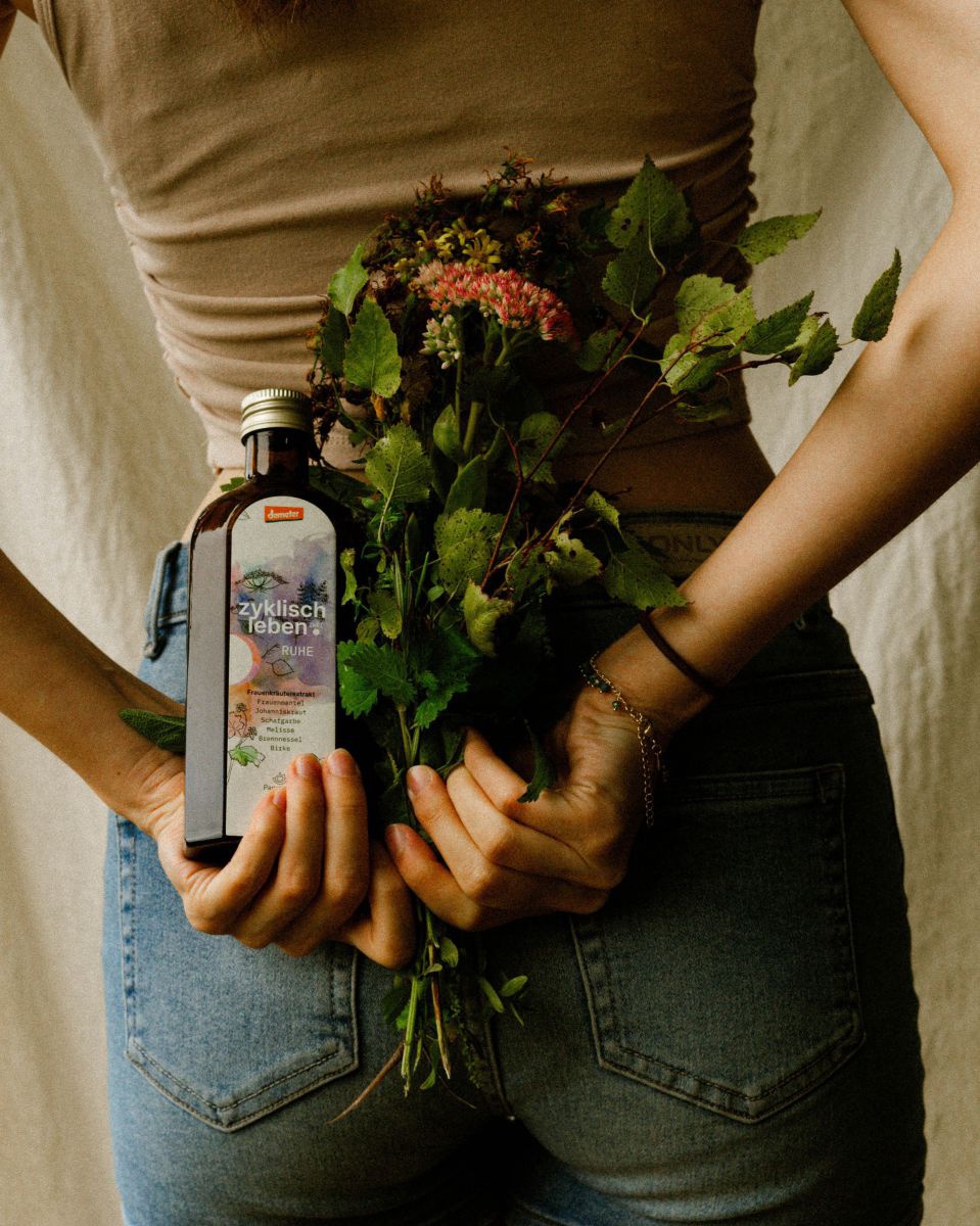 Eine Frau hält hinter ihrem Rücken einen Blumenstrauß und eine Flasche 