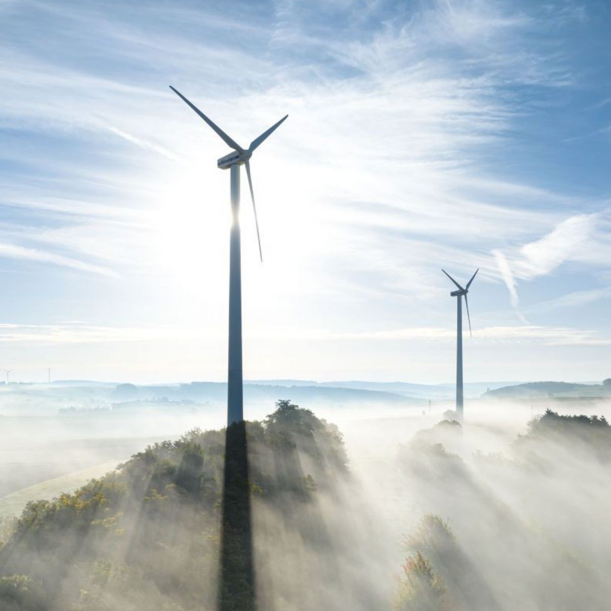 Das Foto zeigt zwei Windräder, die sehr hoch wirken, da die Landschaft darunter von Nebel bedeckt ist. Der Himmel ist weitgehend strahlend blau mit weißen Federwolken durchzogen, alles ist strahlend hell vom Sonnenlicht beleuchtet.