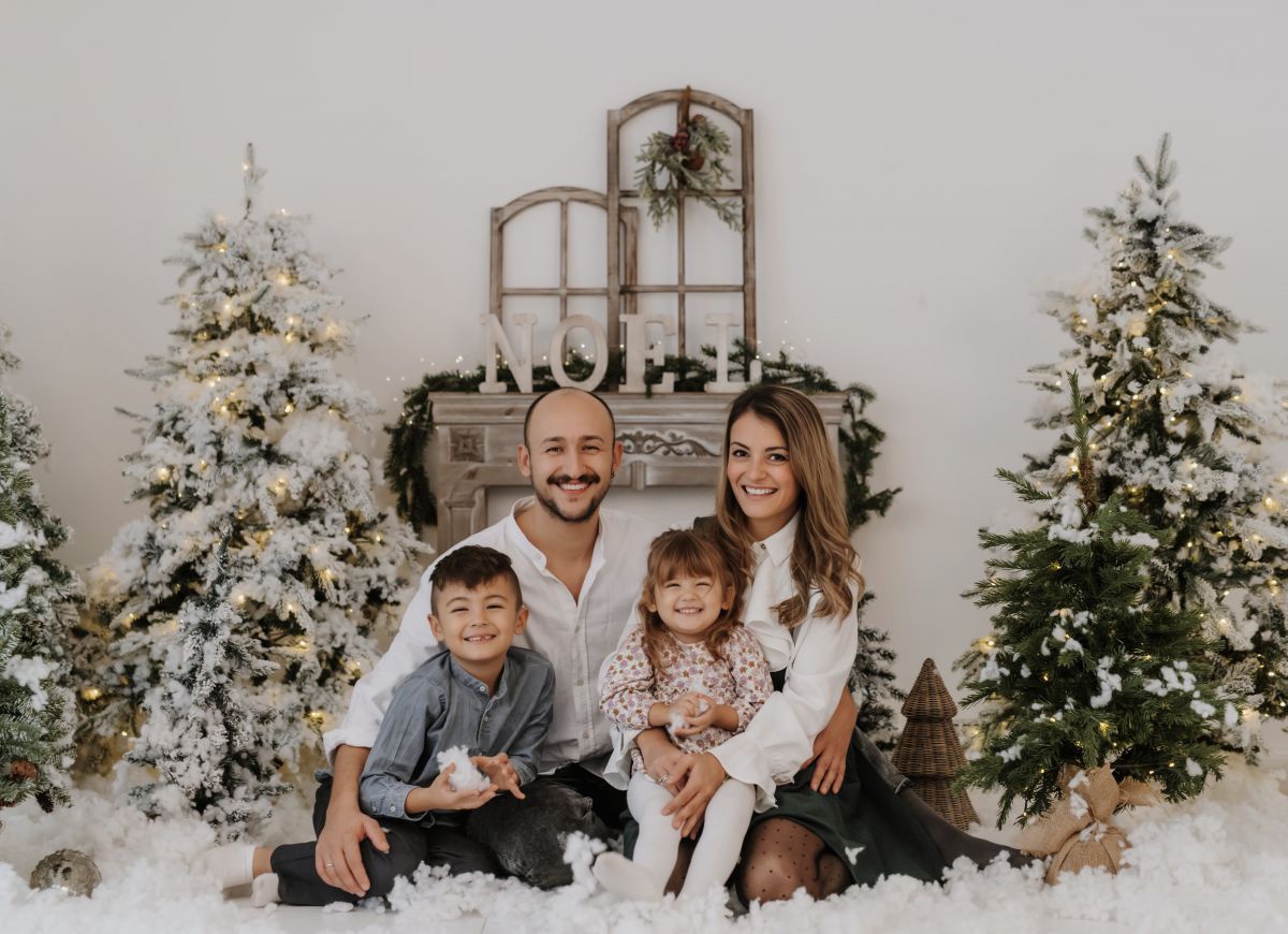 Familie Zusag: Papa Patrik und Mama Jasinta mit ihren Kindern Livi und Kian auf dem Schoß. Die 4 sitzen zusammen am Boden zwischen künstlich beschneiten und beleuchteten Christbäumen und weiterer weihnachtlicher Deko in kühlen Weiß-, Gold- und Brauntönen.