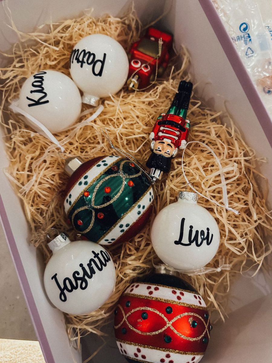 In einer Schuhschachtel liegen auf Holzwolle Christbaumkugeln, 4 davon sind weiß und tragen einen Namen in schwarzer Schrift.