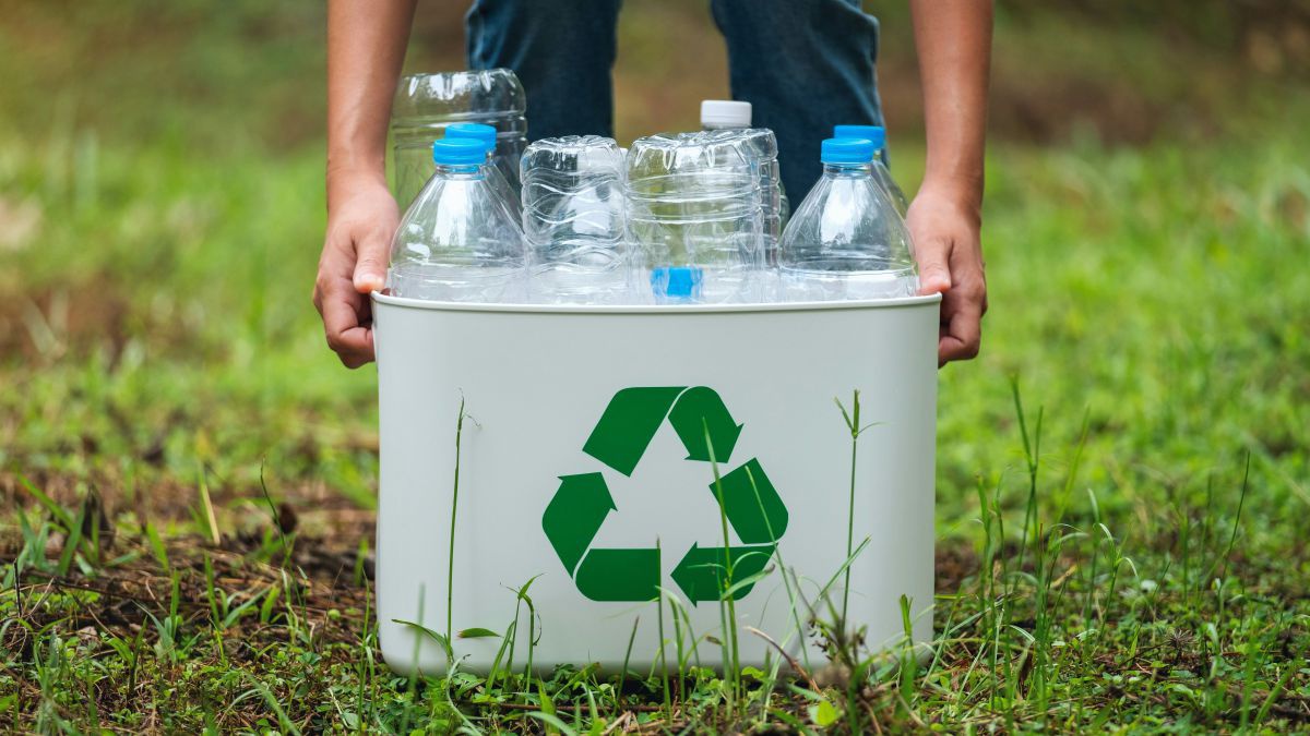 Eine Person hebt einen Kübel mit Plastikflaschen hoch. Auf dem Kübel ist ein Recycling-Logo angebracht.